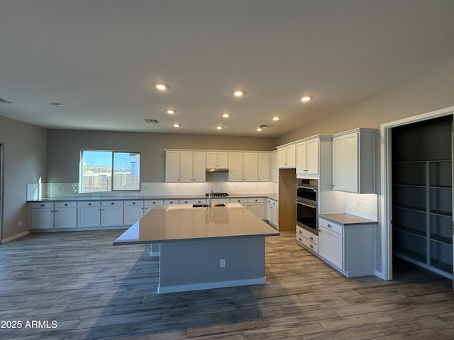 kitchen with a center island with sink, double oven, white cabinetry, and light hardwood / wood-style flooring