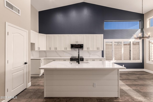 kitchen with hanging light fixtures, high vaulted ceiling, light stone counters, an island with sink, and white cabinets