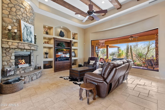 living room featuring a towering ceiling, built in shelves, beam ceiling, ceiling fan, and a stone fireplace