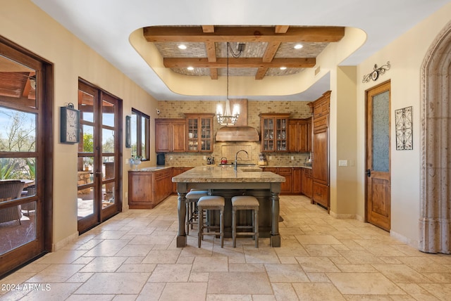 kitchen with an island with sink, light stone countertops, premium range hood, a kitchen bar, and decorative light fixtures