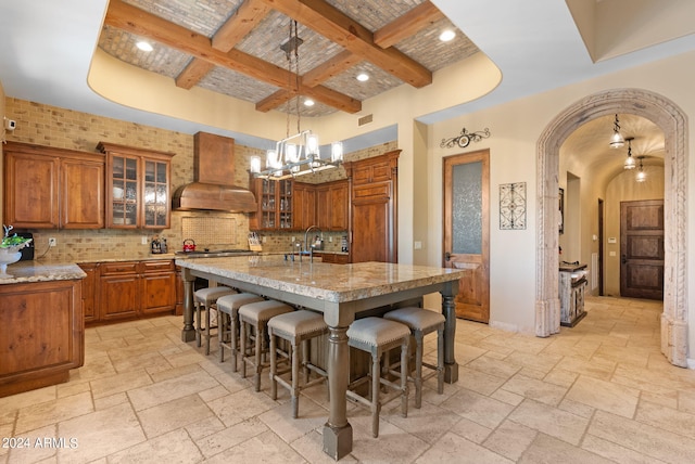 kitchen featuring coffered ceiling, premium range hood, pendant lighting, a kitchen island with sink, and beamed ceiling