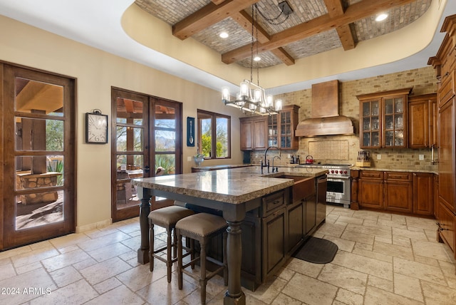 kitchen with a center island with sink, stainless steel range, premium range hood, beam ceiling, and sink