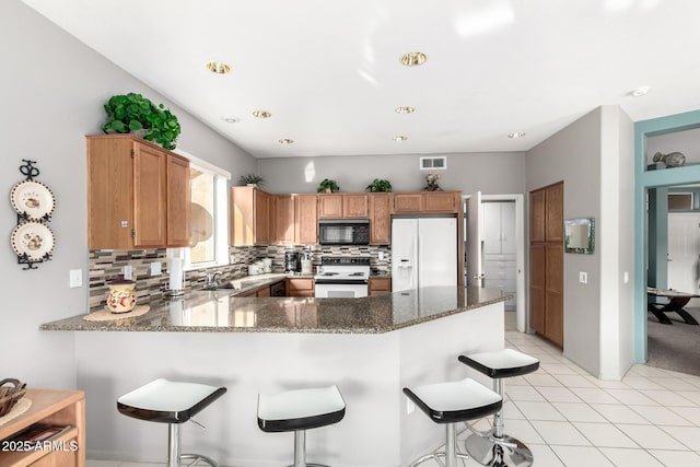 kitchen featuring kitchen peninsula, decorative backsplash, a breakfast bar, white appliances, and dark stone countertops