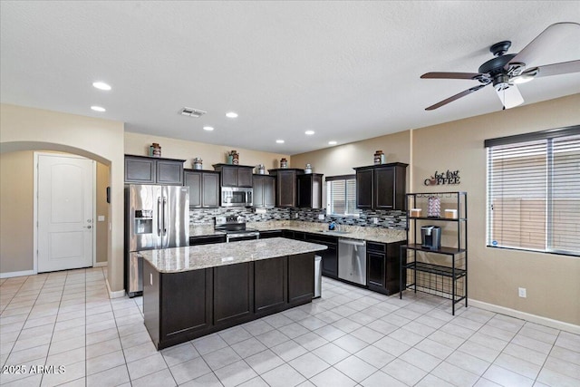 kitchen with light stone counters, arched walkways, appliances with stainless steel finishes, backsplash, and a center island