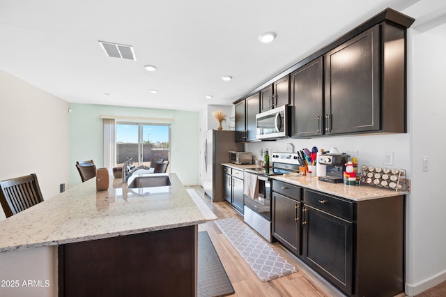 kitchen featuring appliances with stainless steel finishes, sink, light stone counters, a center island with sink, and light hardwood / wood-style flooring