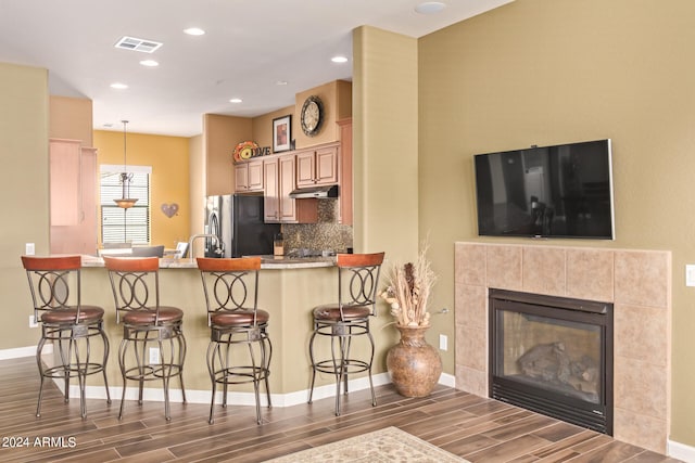 kitchen featuring dark hardwood / wood-style floors, a tiled fireplace, kitchen peninsula, stainless steel fridge, and a kitchen bar