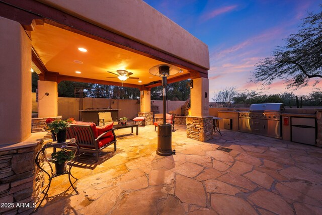 patio terrace at dusk with area for grilling, an outdoor living space, ceiling fan, and exterior kitchen