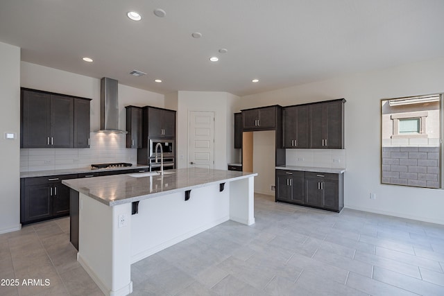kitchen with wall chimney range hood, a kitchen breakfast bar, a sink, a center island with sink, and backsplash