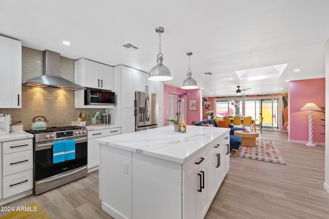 kitchen with wall chimney exhaust hood, a skylight, appliances with stainless steel finishes, a kitchen island, and white cabinets