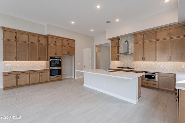 kitchen with decorative backsplash, appliances with stainless steel finishes, wall chimney exhaust hood, a kitchen island with sink, and sink