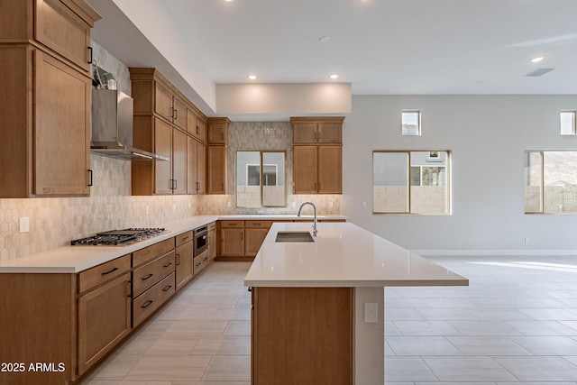 kitchen with appliances with stainless steel finishes, backsplash, sink, wall chimney range hood, and a center island with sink