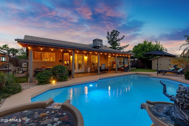 pool at dusk featuring an outdoor hot tub, a fenced backyard, a shed, a patio area, and an outbuilding