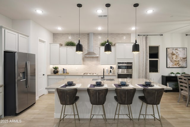 kitchen featuring sink, stainless steel appliances, wall chimney range hood, a center island with sink, and white cabinets