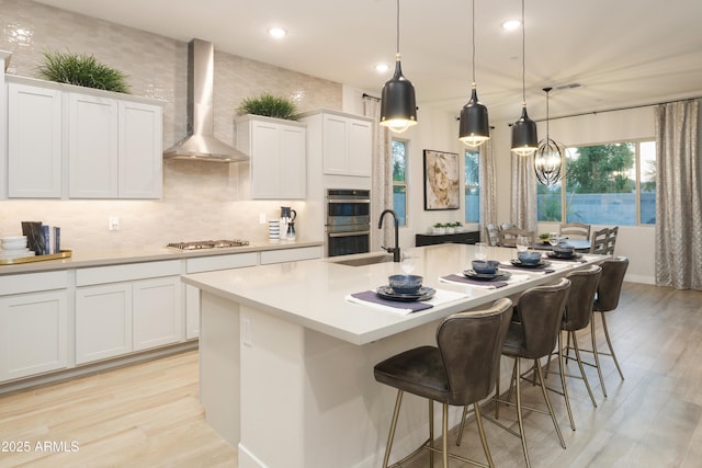 kitchen featuring stainless steel appliances, a kitchen island with sink, wall chimney range hood, decorative light fixtures, and white cabinetry