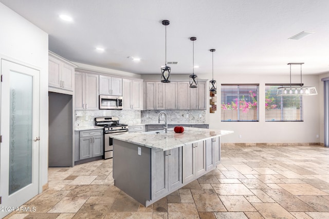 kitchen featuring gray cabinetry, appliances with stainless steel finishes, hanging light fixtures, light stone counters, and a center island with sink