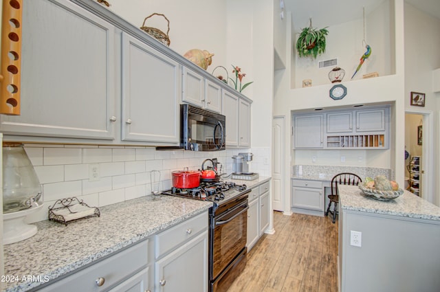 kitchen with light stone countertops, light wood-type flooring, black appliances, gray cabinetry, and a towering ceiling