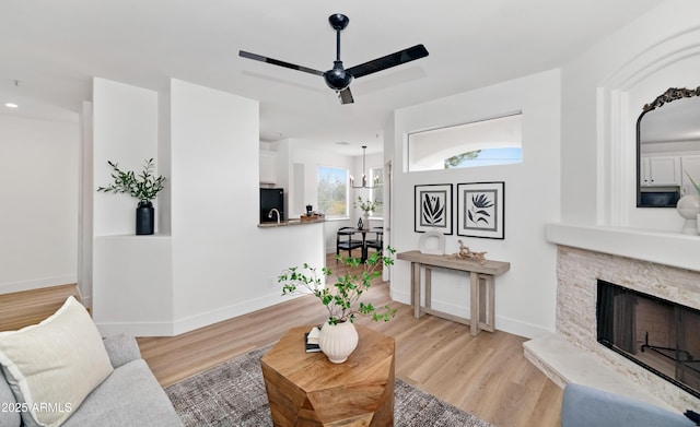 living area featuring light wood-style floors, ceiling fan, a stone fireplace, and baseboards