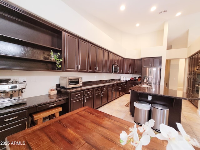 kitchen featuring dark brown cabinets, appliances with stainless steel finishes, dark stone counters, light tile patterned floors, and a toaster