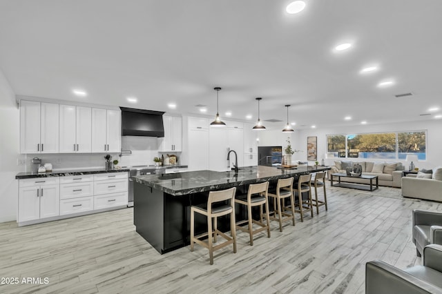 kitchen featuring white cabinetry, decorative light fixtures, dark stone countertops, a large island with sink, and wall chimney range hood