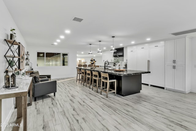 kitchen featuring white cabinetry, light hardwood / wood-style flooring, hanging light fixtures, a center island with sink, and dark stone counters