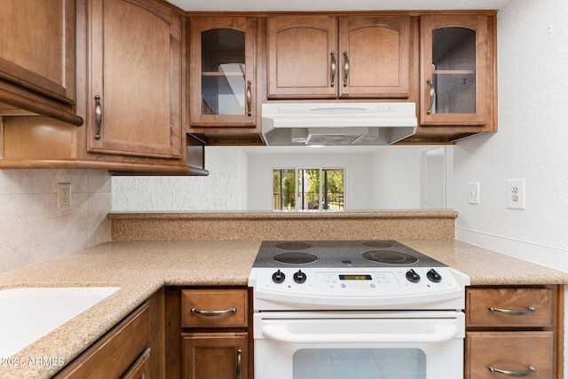 kitchen with under cabinet range hood, white electric range, backsplash, brown cabinetry, and glass insert cabinets