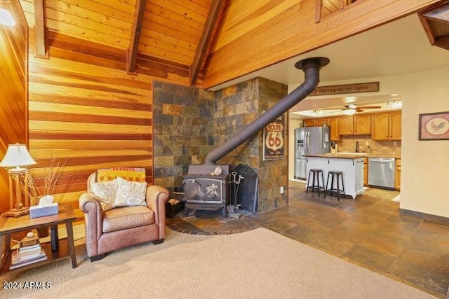 living room featuring beam ceiling, a wood stove, ceiling fan, wood walls, and wood ceiling