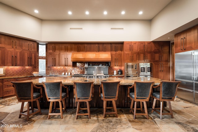 kitchen featuring a towering ceiling, light tile flooring, tasteful backsplash, a large island with sink, and built in appliances