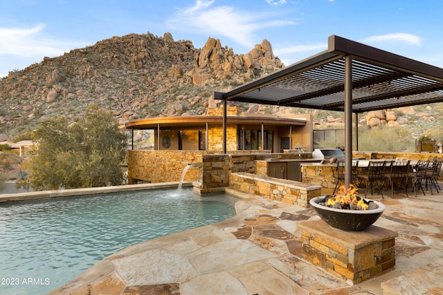 view of pool featuring a grill, pool water feature, an outdoor kitchen, a mountain view, and a pergola