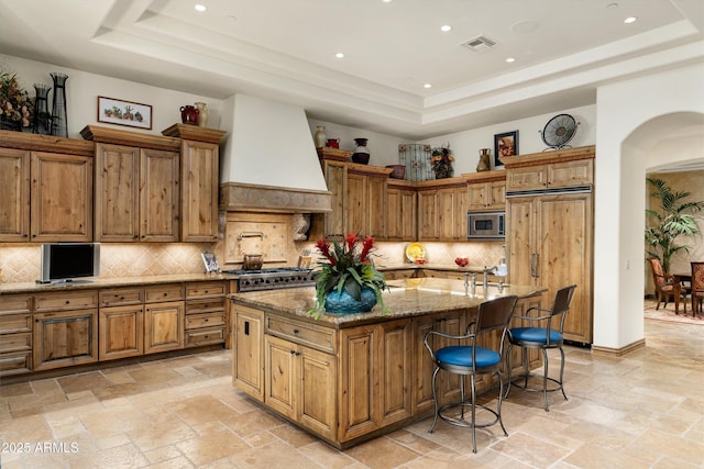 kitchen featuring a kitchen island with sink, built in appliances, light stone counters, a tray ceiling, and custom range hood