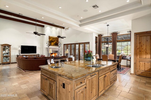 kitchen with a raised ceiling, an island with sink, sink, and light stone counters