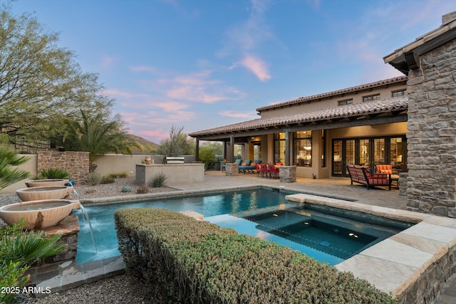 pool at dusk featuring pool water feature, an outdoor kitchen, outdoor lounge area, an in ground hot tub, and a patio