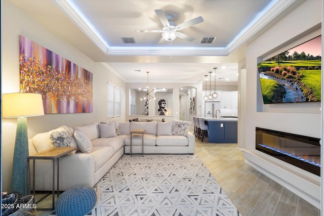 living room with visible vents, a tray ceiling, light wood-style floors, crown molding, and ceiling fan with notable chandelier