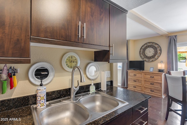 kitchen featuring wood finished floors, a sink, dark brown cabinets, dark countertops, and beamed ceiling