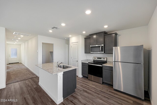 kitchen with light stone counters, a kitchen island with sink, dark colored carpet, appliances with stainless steel finishes, and sink