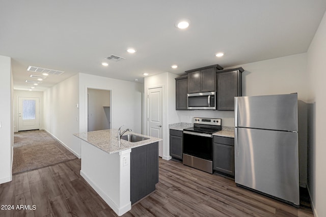 kitchen with dark wood-type flooring, sink, light stone counters, an island with sink, and stainless steel appliances