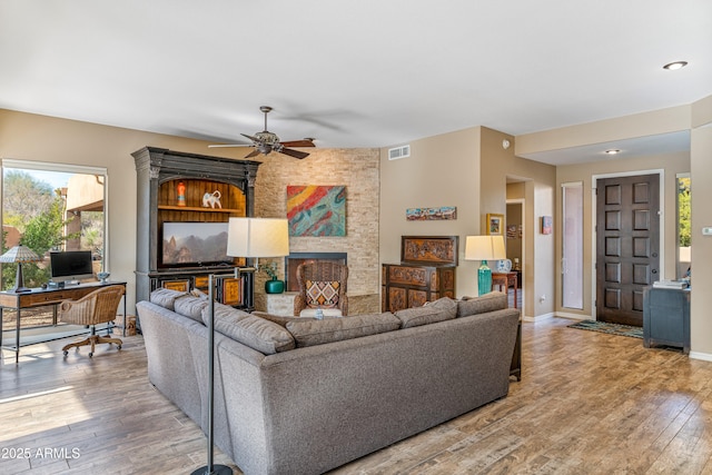 living room featuring a stone fireplace, wood-type flooring, and ceiling fan