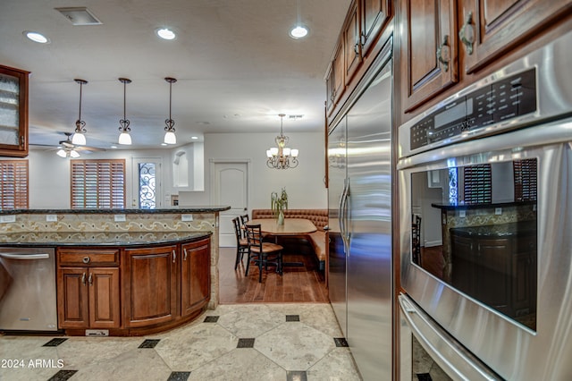 kitchen with ceiling fan with notable chandelier, dark stone counters, tile floors, stainless steel appliances, and pendant lighting