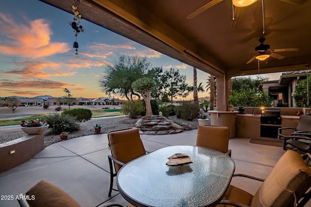 patio terrace at dusk featuring an outdoor kitchen, a ceiling fan, a residential view, grilling area, and outdoor dining area