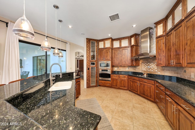 kitchen featuring wall chimney range hood, appliances with stainless steel finishes, dark stone counters, pendant lighting, and sink