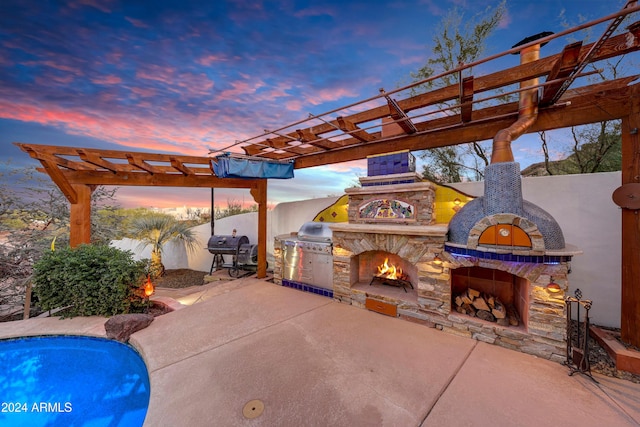 patio terrace at dusk with an outdoor stone fireplace, grilling area, and a pergola