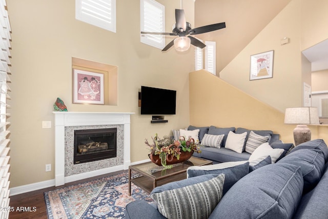 living area with a ceiling fan, a glass covered fireplace, dark wood finished floors, baseboards, and a towering ceiling