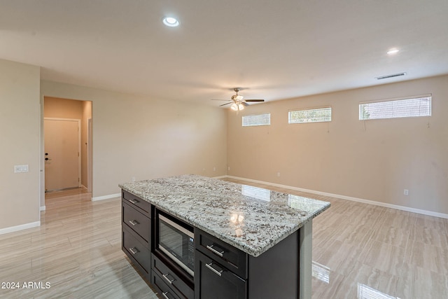 kitchen with stainless steel microwave, a center island, light hardwood / wood-style floors, ceiling fan, and light stone counters