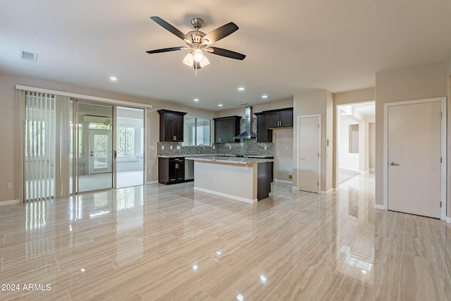 kitchen featuring light stone countertops, wall chimney range hood, backsplash, a center island, and ceiling fan