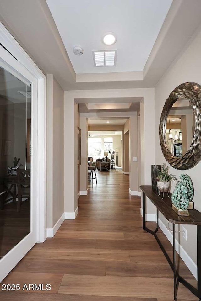 corridor featuring a raised ceiling, wood finished floors, visible vents, and baseboards