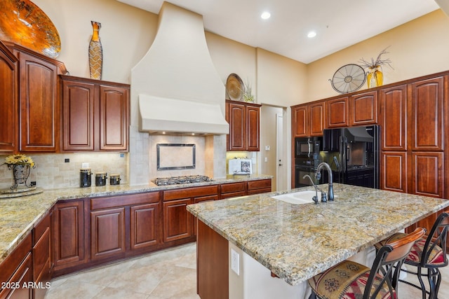 kitchen with light stone countertops, custom range hood, a center island with sink, and black appliances