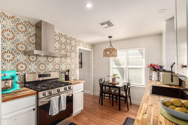 kitchen featuring butcher block counters, wall chimney exhaust hood, hanging light fixtures, stainless steel range with gas cooktop, and white cabinets