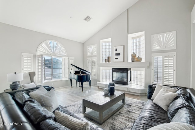 living area featuring high vaulted ceiling, light wood-style flooring, visible vents, and a glass covered fireplace