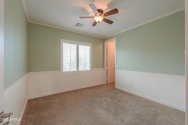 carpeted spare room with ornamental molding, wainscoting, visible vents, and a ceiling fan