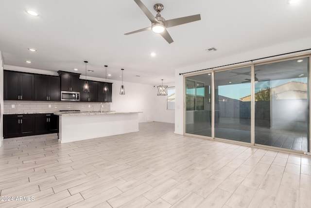 kitchen featuring light stone counters, pendant lighting, ceiling fan with notable chandelier, decorative backsplash, and a kitchen island with sink