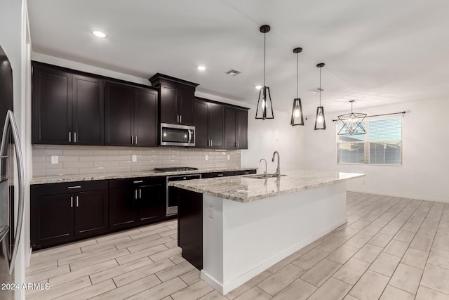 kitchen with a center island with sink, stainless steel appliances, light stone countertops, sink, and decorative light fixtures